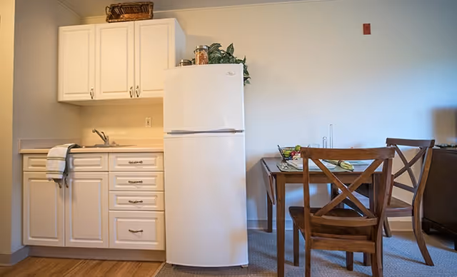 A small kitchen area with white cabinets, a countertop with a sink, and a white refrigerator. Next to the kitchen is a wooden dining table with two wooden chairs. The table has a placemat, a bowl of fruit, and some utensils. The room has light-colored walls and a wooden floor.