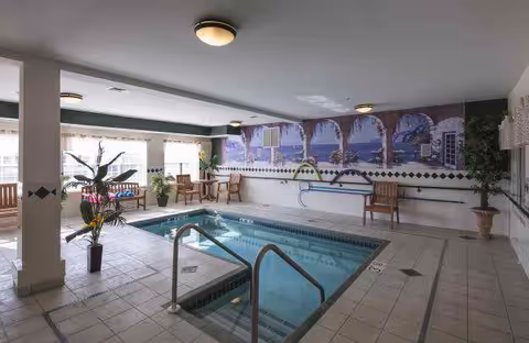 Indoor swimming pool area with tiled floor and walls, featuring a small pool with metal handrails. Several wooden chairs and potted plants are arranged around the pool. A large mural depicting an ocean view with arches and palm trees decorates one wall. The room is lit by ceiling lights and natural light from large windows.