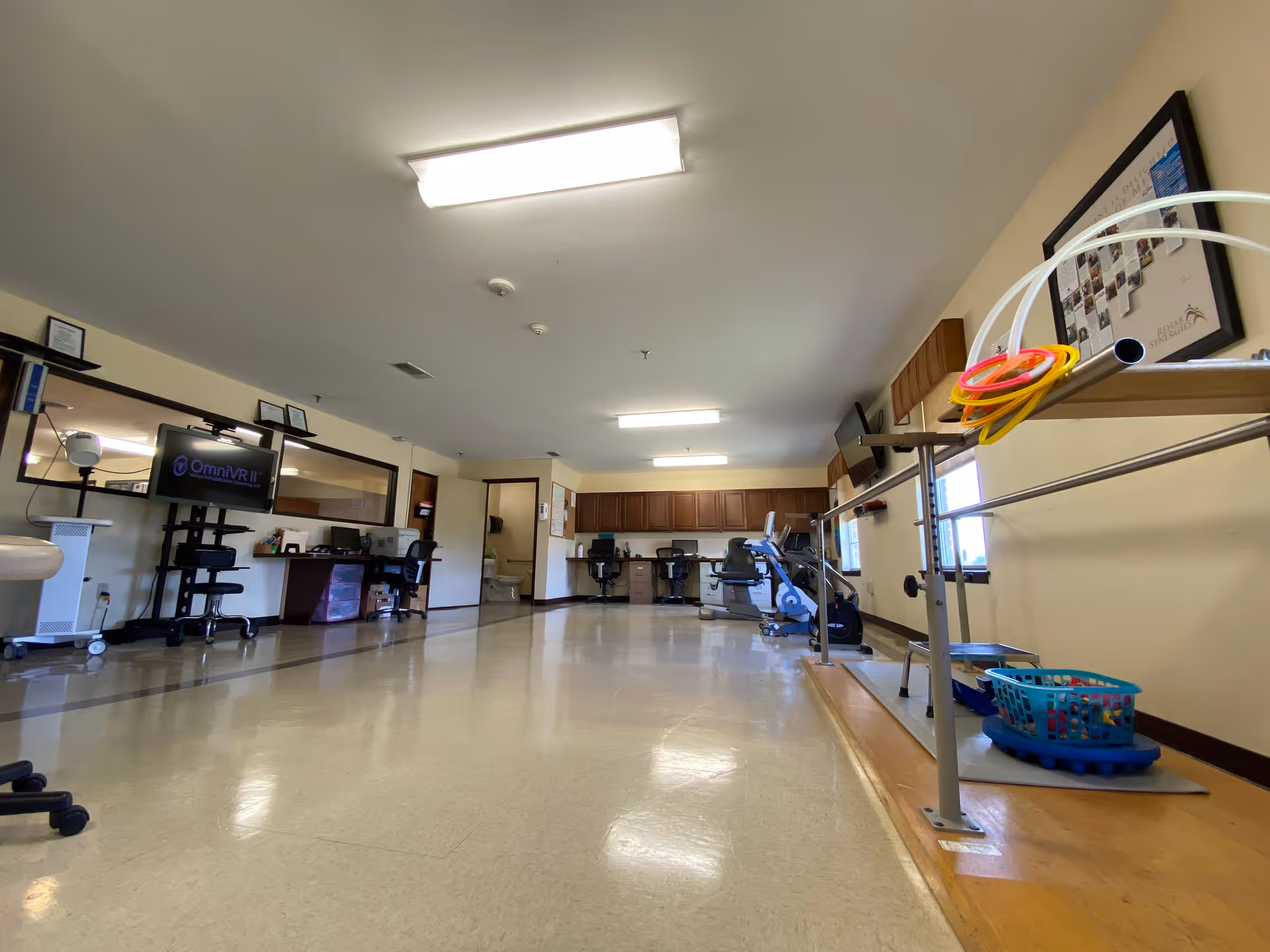 Spacious therapy room in a nursing facility with exercise equipment, parallel bars, desks and monitors.
