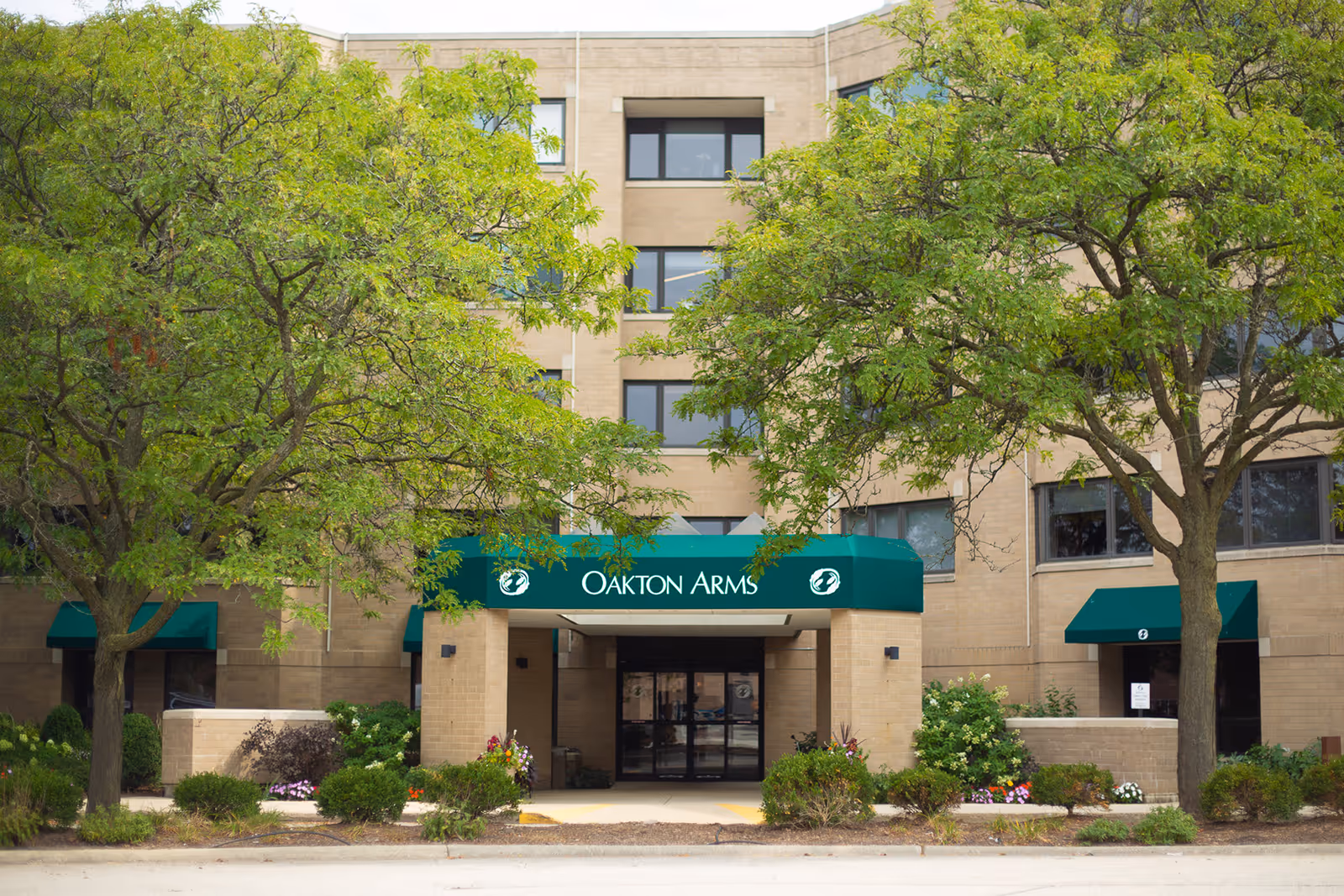 Front exterior view of the Oakton Arms building with a green awning over the entrance, surrounded by trees and landscaping.