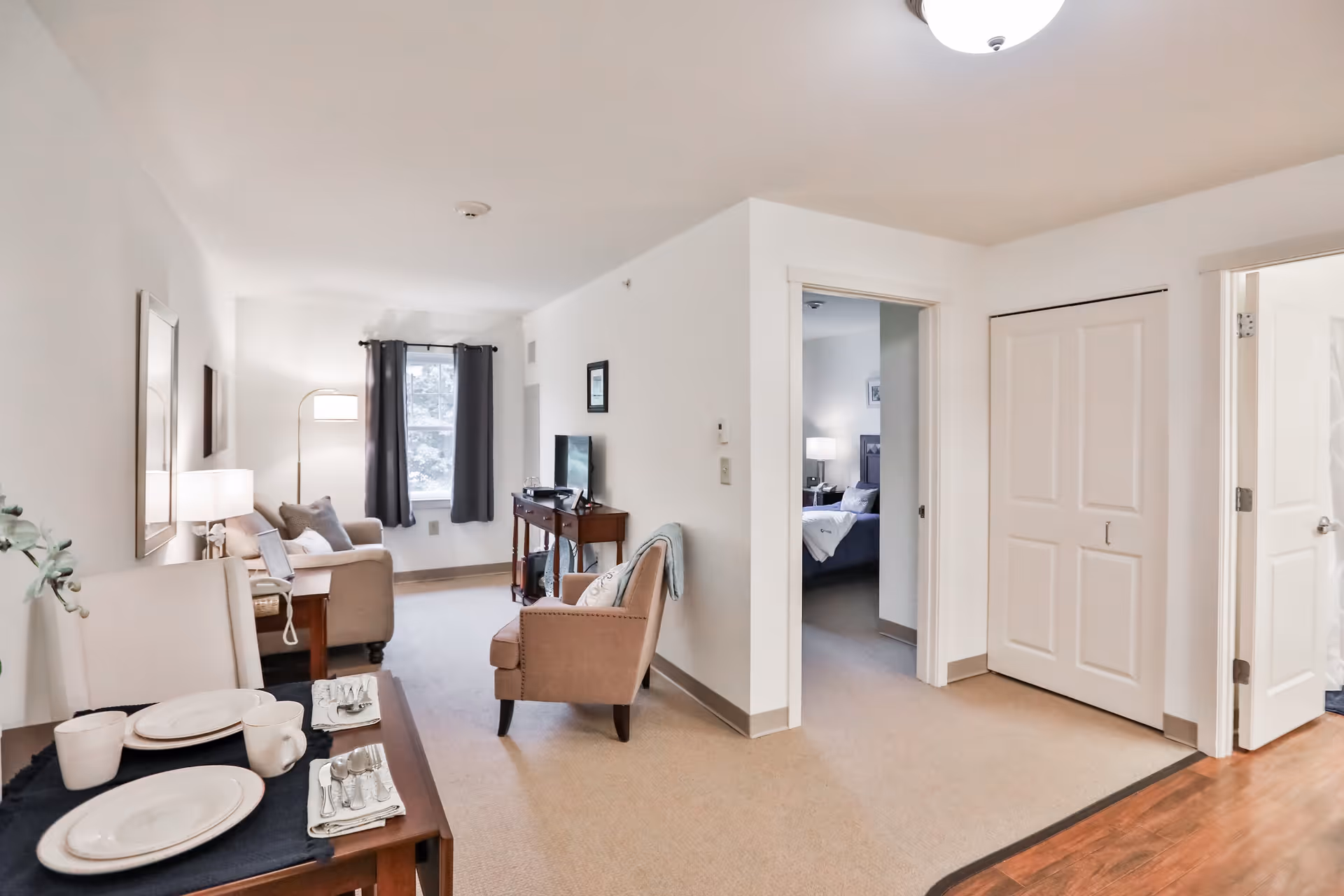 Interior view of a senior living apartment showing a small dining table set with plates, cups, and silverware in the foreground. Beyond the table is a living area with two armchairs, a side table with a lamp, a TV on a console, and a window with dark curtains. To the right, there is a hallway leading to a bedroom with a bed and nightstand visible, and a closed closet door. The walls are white and the flooring is a mix of carpet and wood.