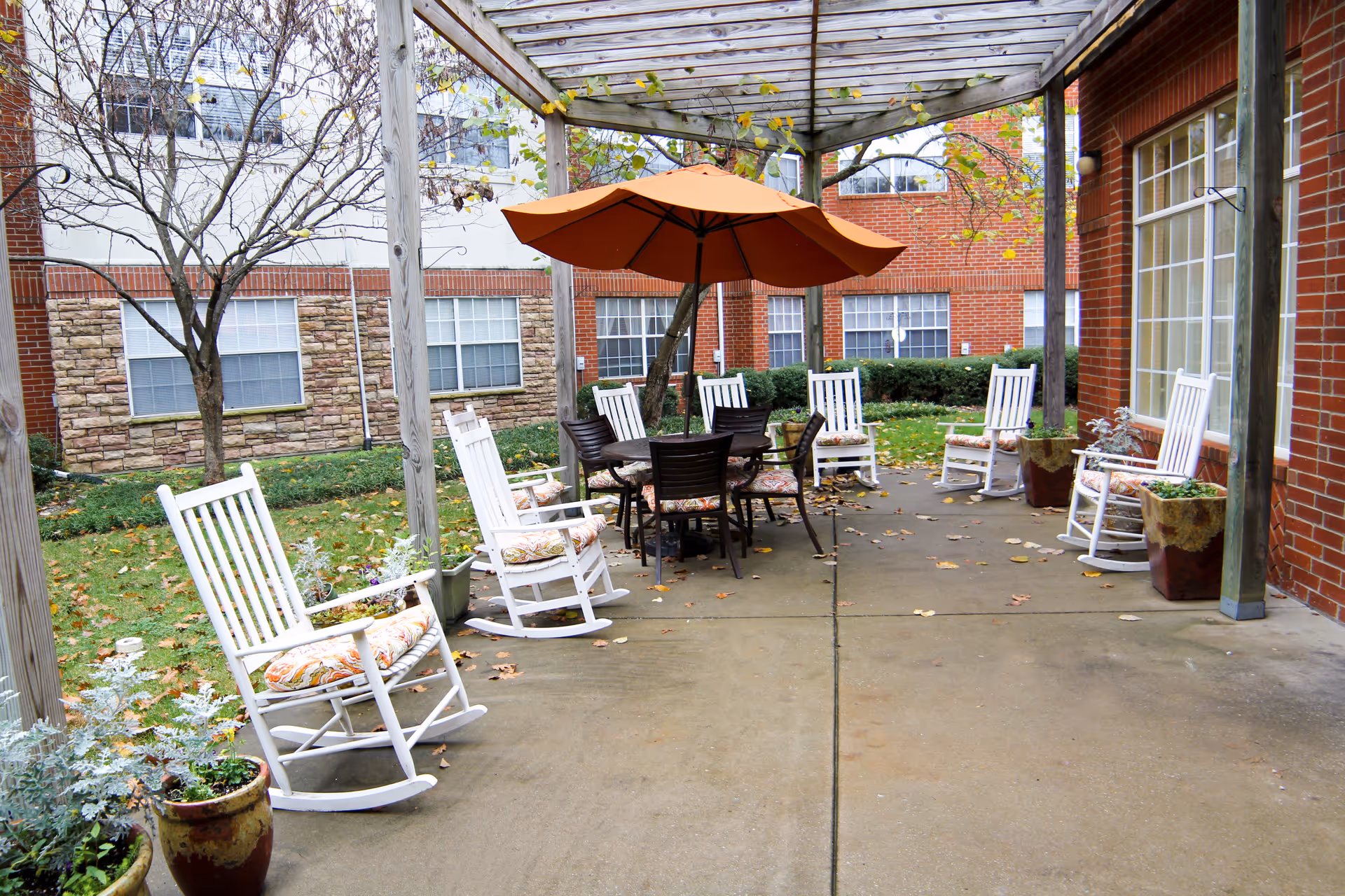 Courtyard patio with white rocking chairs arranged under a pergola around a table with an orange umbrella in front of a brick building.