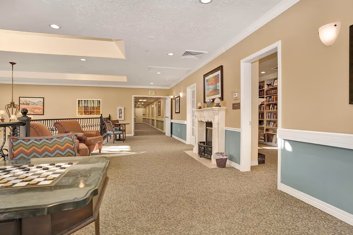 A spacious and well-lit common area in a senior living facility featuring comfortable seating with patterned cushions, a fireplace with decorative items on the mantel, framed artwork on the walls, and a hallway leading to other rooms. There is a chess/checkers table in the foreground and a room with bookshelves visible through an open door.