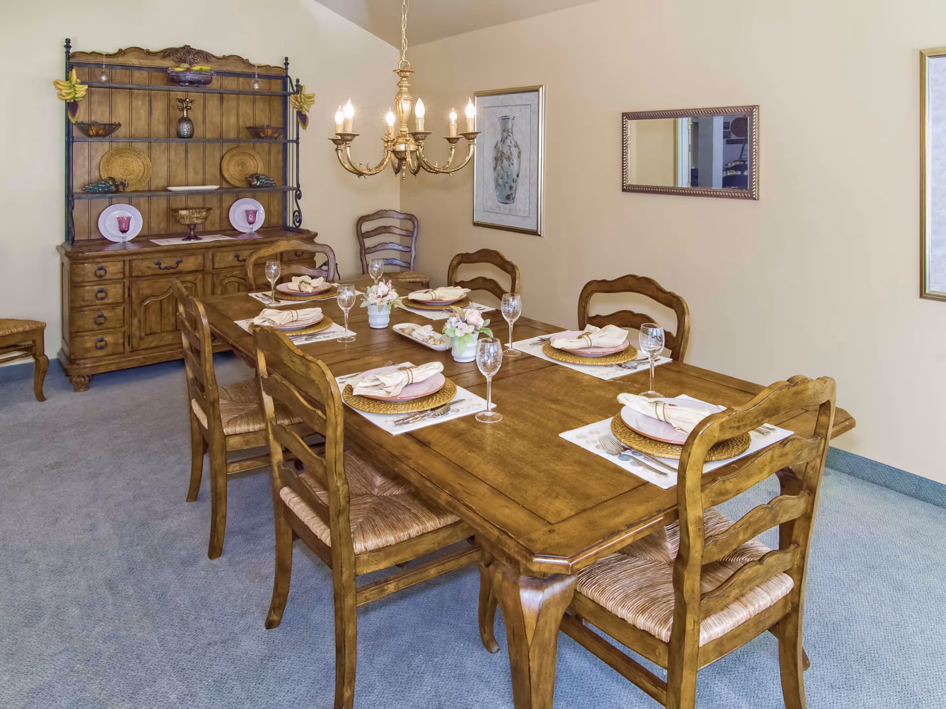 A wooden dining table set for six with place settings, chairs, a chandelier overhead, and a wooden hutch against the wall.