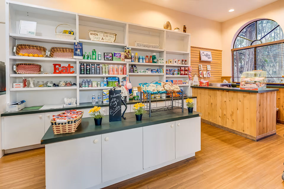 Interior view of a small convenience or snack shop area inside a senior living facility. Shelves are stocked with various snacks, drinks, and personal care items. A counter labeled 'Coffee Bar' is visible to the right with a display case containing baked goods. The space has wooden flooring, white cabinetry, and a large arched window with blinds letting in natural light.