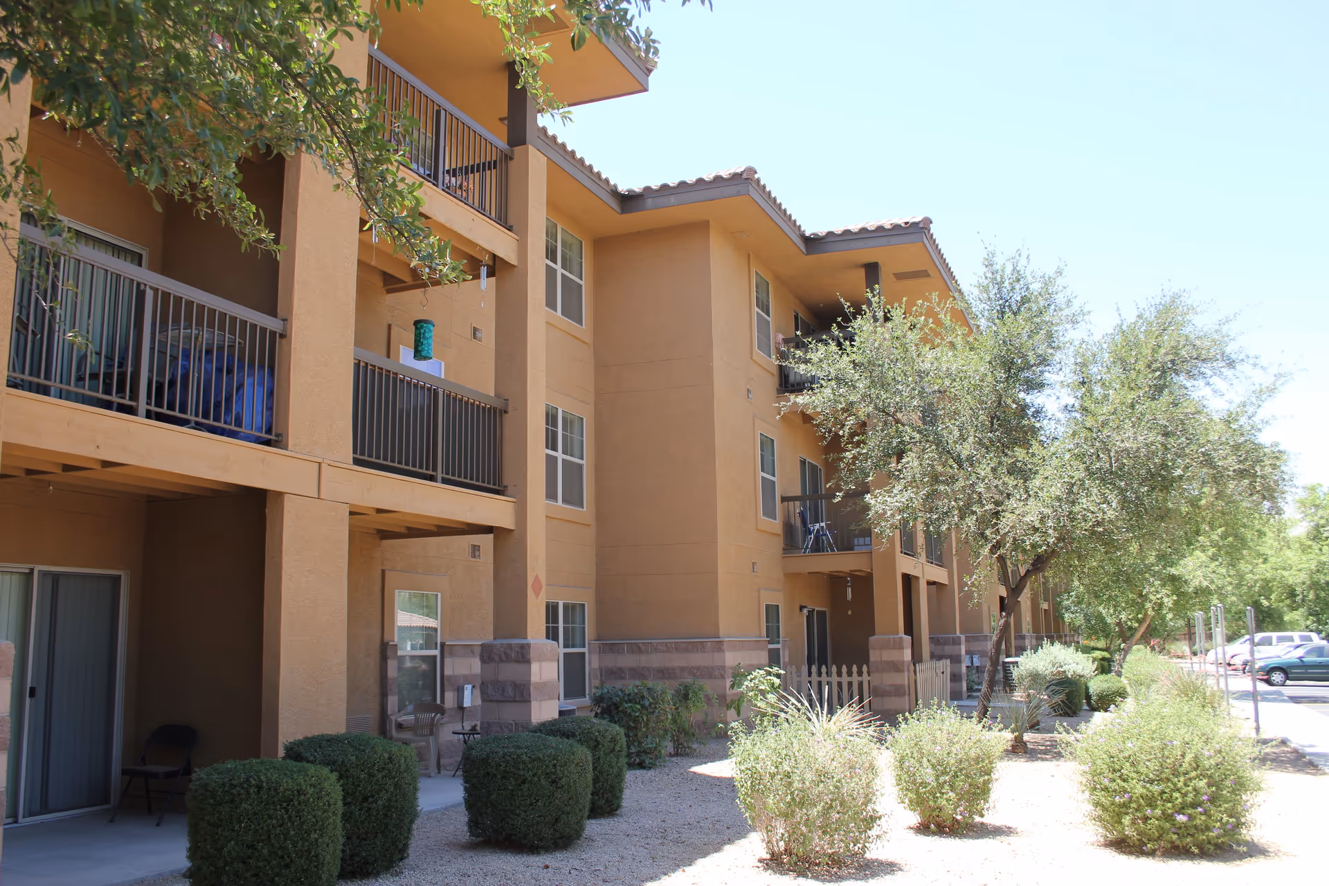 Exterior view of a three-story residential building with balconies, surrounded by trees and shrubs under a clear blue sky.