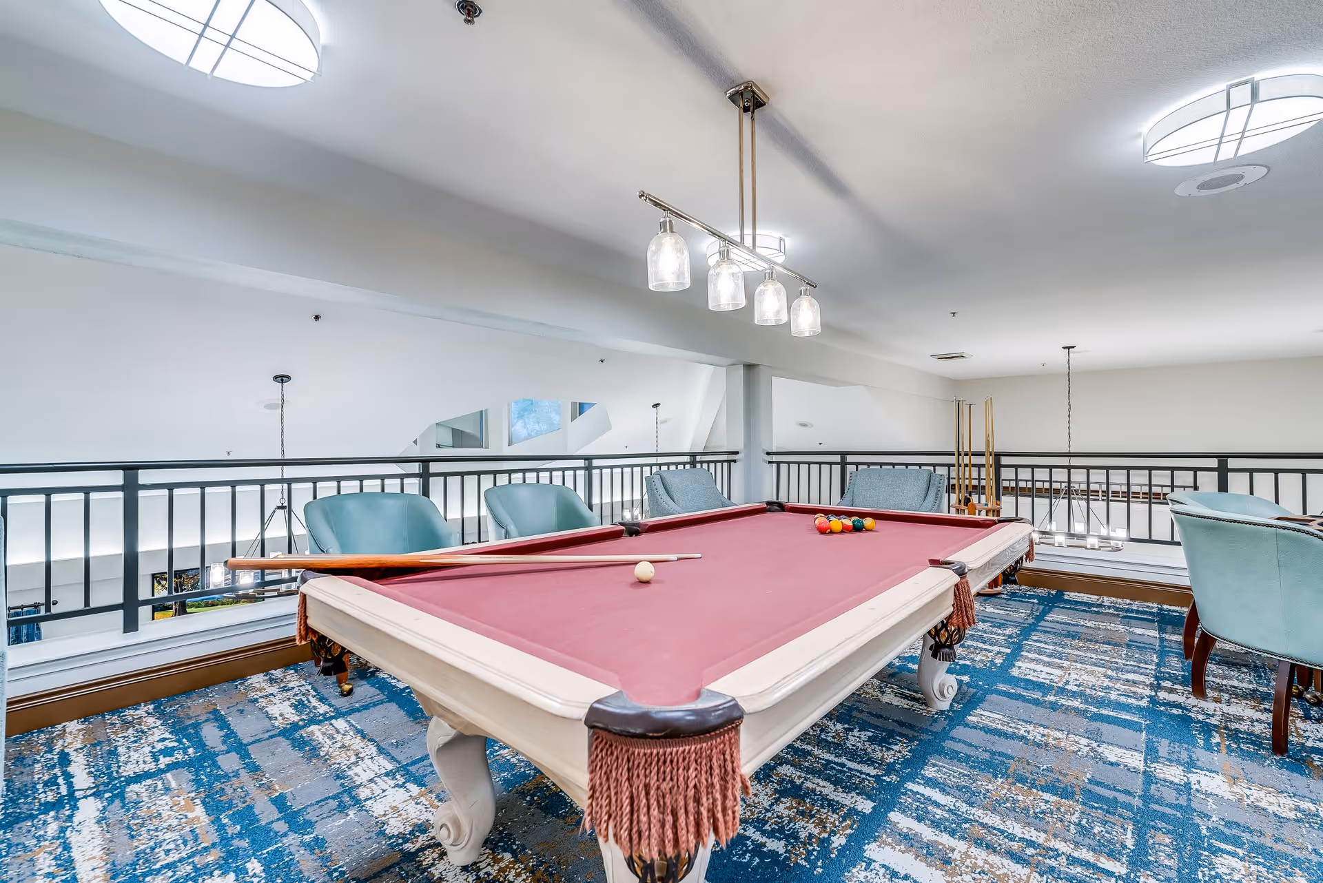 Interior view of a senior living facility's game room featuring a red felt pool table with pool cues and balls arranged on it. The room has blue patterned carpet, teal chairs around the pool table, and modern ceiling lights. A black railing overlooks a lower level.