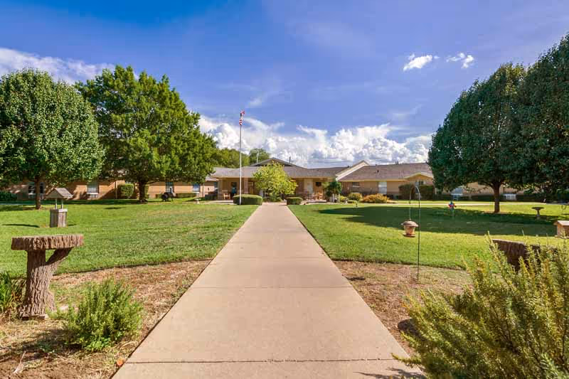 A wide concrete pathway leads through a well-maintained green lawn with trees and shrubs on either side, towards a single-story building under a partly cloudy blue sky. An American flag is visible near the building entrance.
