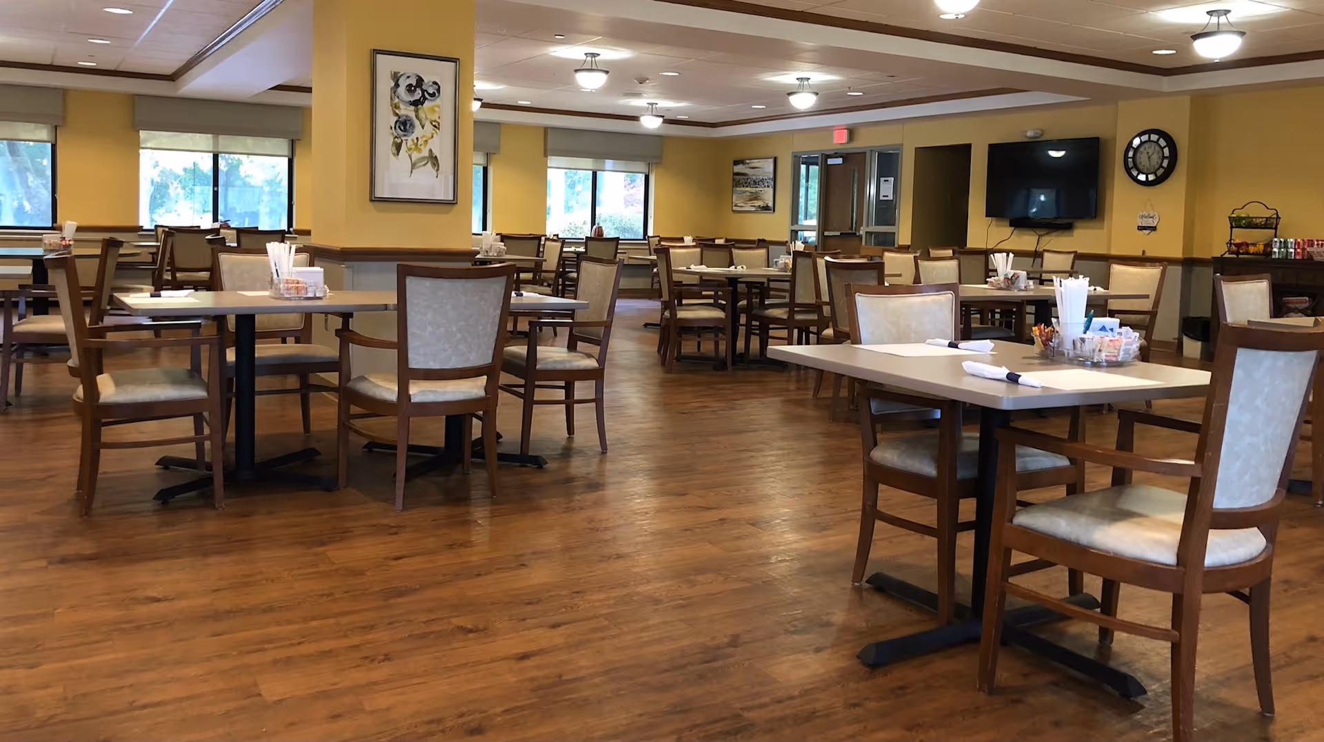 A spacious dining room in a senior living facility with multiple wooden tables and chairs arranged neatly. The room has large windows letting in natural light, yellow walls, wooden flooring, and ceiling lights. There is a wall-mounted TV, a clock, and some artwork on the walls.