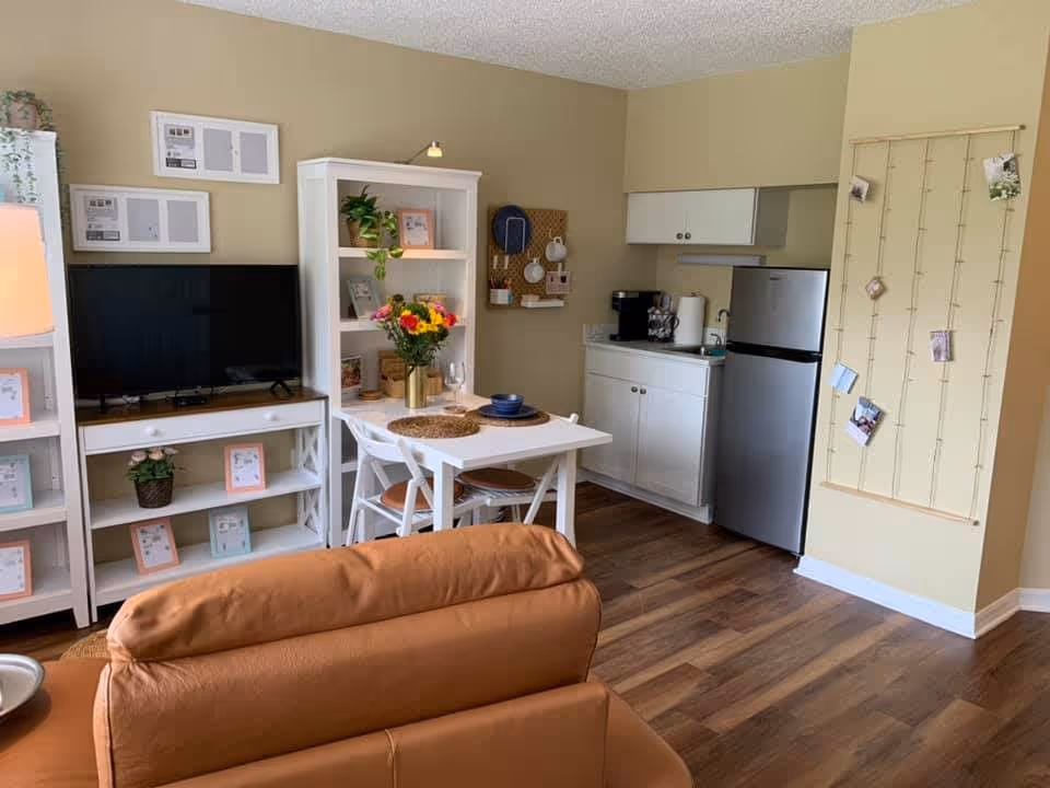A cozy living area with a brown leather sofa in the foreground, a white dining table with two chairs set with placemats and dishes, a white bookshelf with plants and picture frames, a flat-screen TV on a white stand, and a small kitchen area with white cabinets, a stainless steel refrigerator, and a coffee maker. The walls are beige and the floor is wood.