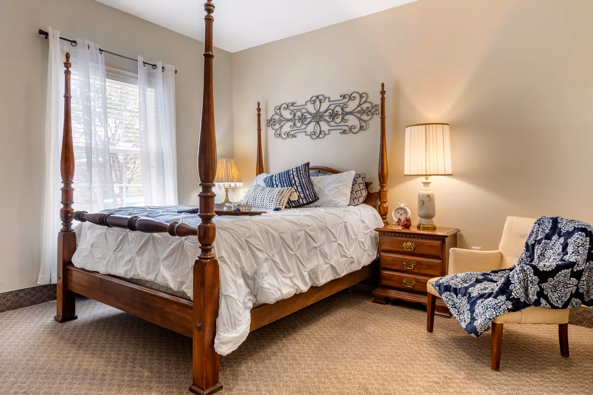 A cozy bedroom with a wooden four-poster bed dressed in white bedding and multiple decorative pillows. Next to the bed is a wooden nightstand with a lamp, an alarm clock, and a small decorative item. A beige armchair with a blue patterned throw blanket is positioned to the right of the nightstand. Sheer white curtains cover a window on the left side of the room, and a decorative metal wall art piece hangs above the bed.