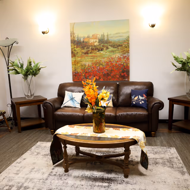 A cozy living room with a brown leather sofa adorned with three decorative pillows. In front of the sofa is a wooden coffee table with a floral table runner and a vase of bright orange and yellow flowers. On either side of the sofa are wooden side tables with glass vases containing green leafy plants. A large colorful landscape painting hangs on the wall above the sofa, and two wall-mounted lights illuminate the space. The floor is covered with a light-colored patterned rug.