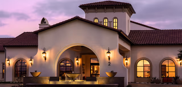 Exterior view of a cream-colored building with arches, warm lights, and a fountain at the entrance.