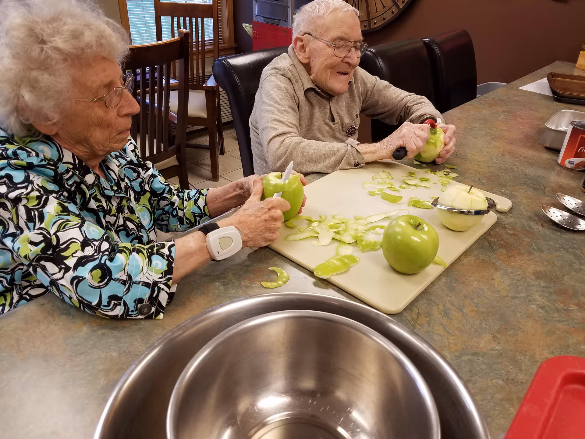 Two elderly individuals sitting at a kitchen counter peeling green apples. One person is wearing a patterned shirt and a medical alert bracelet, while the other is smiling and wearing glasses. There are apple peels and kitchen utensils on the counter.