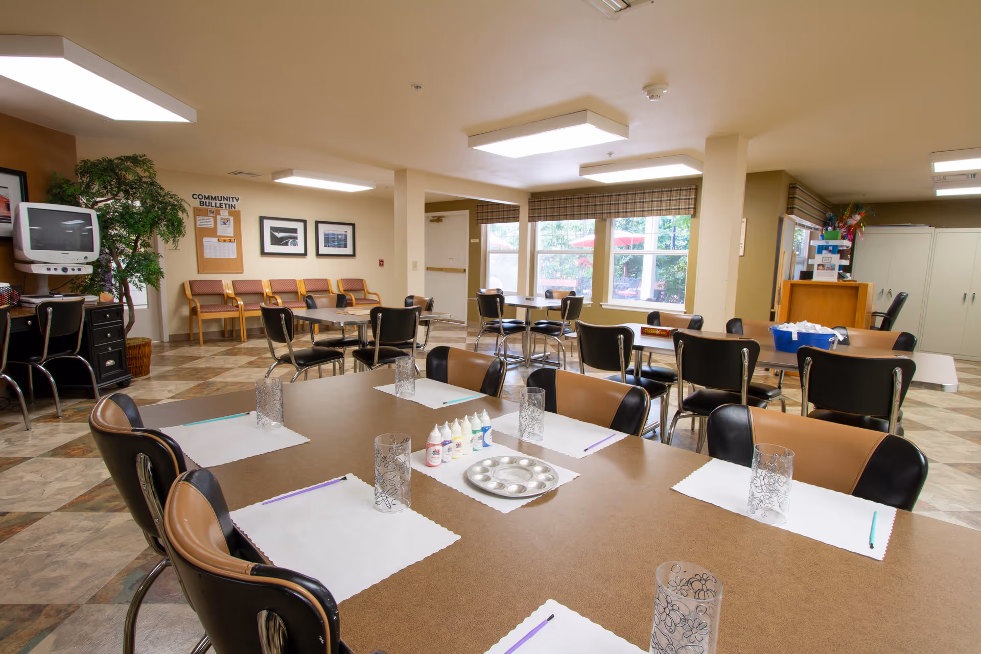 A spacious common room in a senior living facility with multiple tables and chairs arranged for group activities. The tables have placemats, glasses, and art supplies such as paint bottles and brushes. The room has large windows with a view of an outdoor patio area, a community bulletin board, framed pictures on the wall, and a TV on a stand. The floor is tiled, and the room is well-lit with ceiling lights.