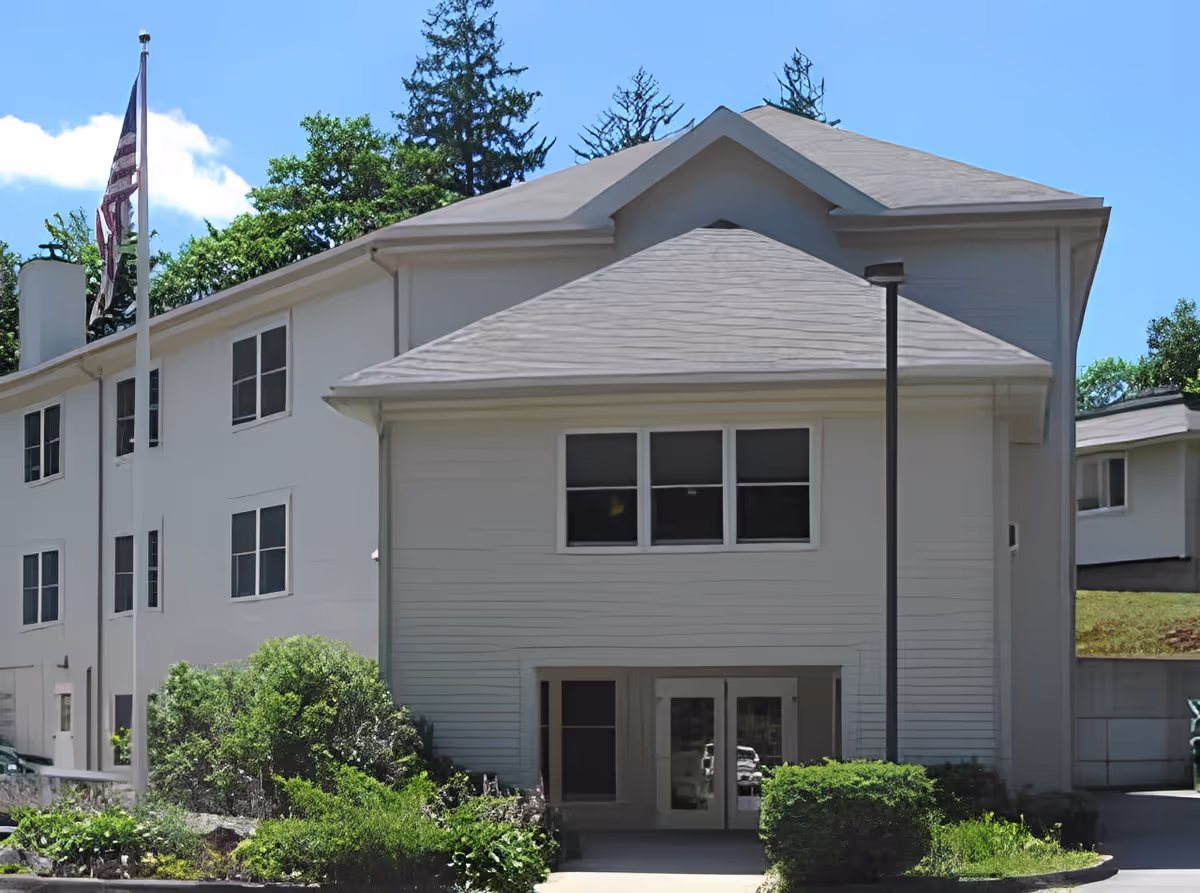 Exterior view of a two-story white building with multiple windows, an American flag on a flagpole, and surrounding greenery under a clear blue sky.