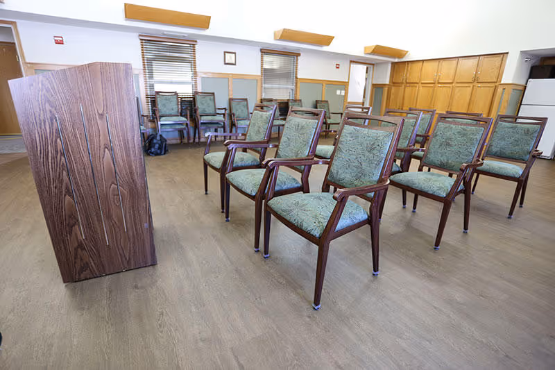 A room with several wooden chairs with green patterned cushions arranged in rows facing a wooden podium. The room has wood flooring, windows with blinds, and wooden paneling on one wall.