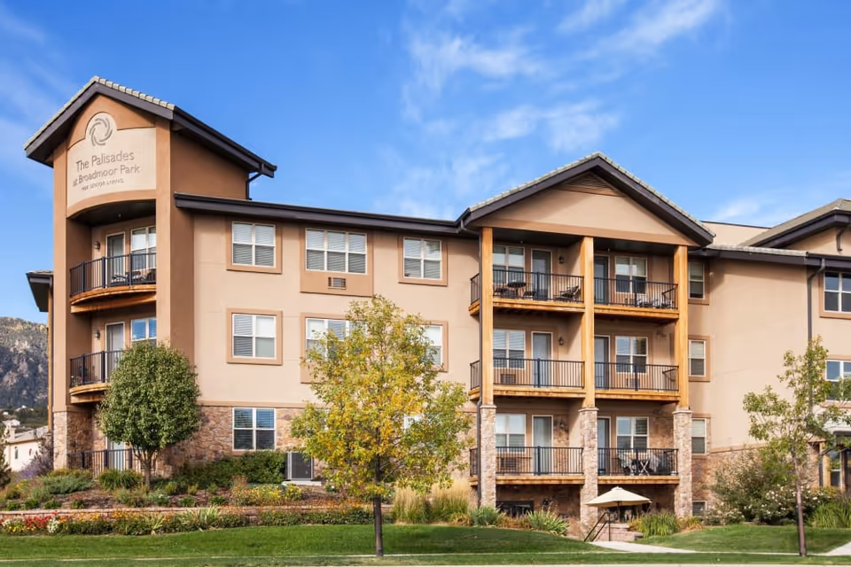 Exterior view of The Palisades at Broadmoor Park senior living facility, showing a multi-story building with balconies, stone and beige stucco facade, surrounded by landscaped greenery and trees under a blue sky.