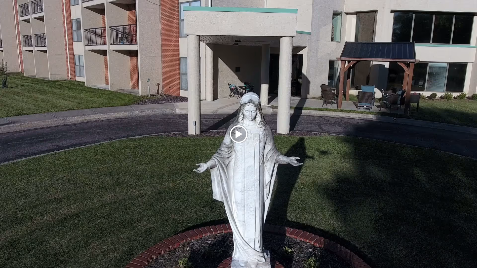 Outdoor view of a senior living facility with a white statue of Our Lady of Mercy standing on a circular brick-bordered patch of grass in front of the building entrance. The building has balconies and a covered seating area with chairs and a small roofed structure on the right side.