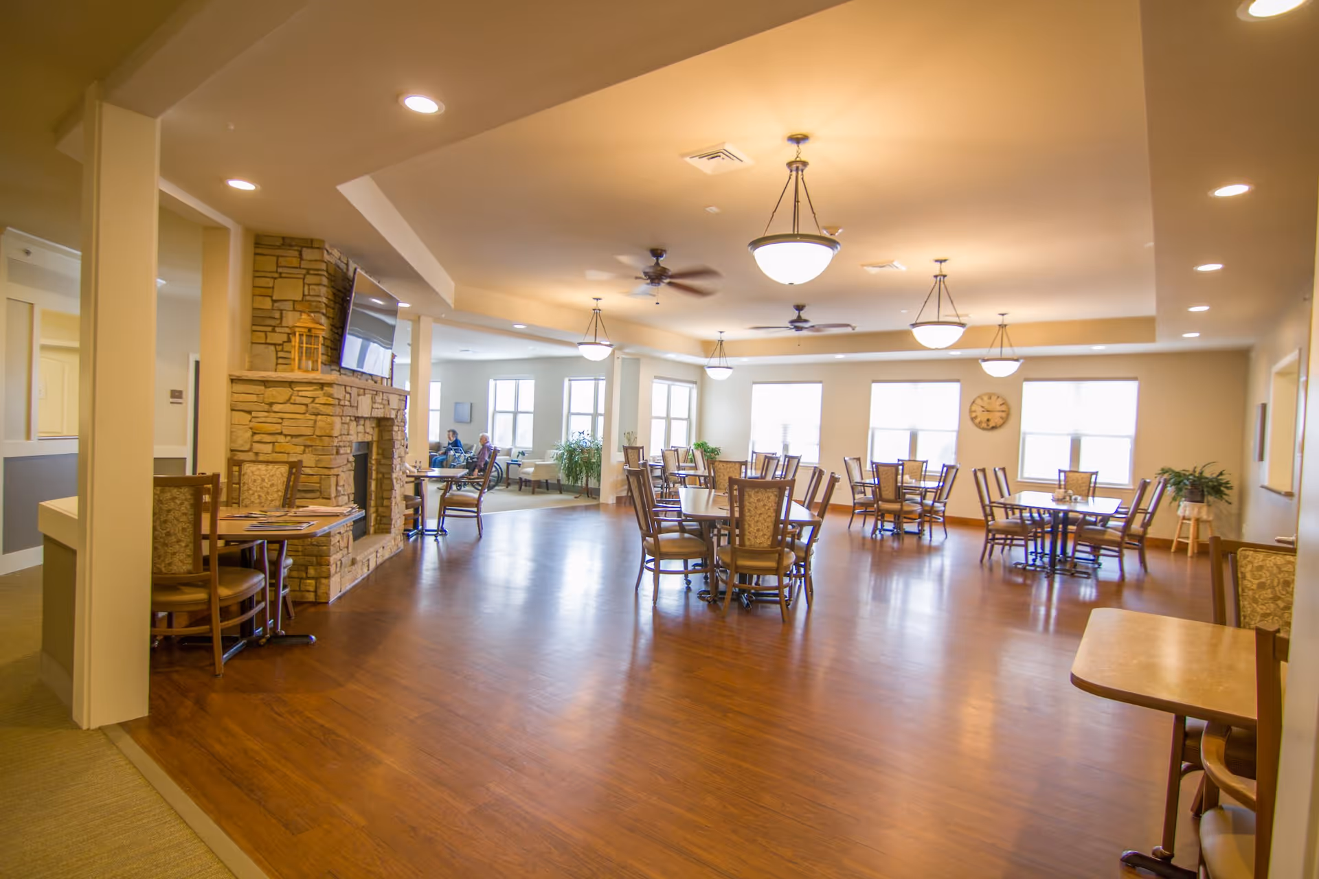 A spacious dining room in an assisted living facility with multiple round tables and chairs arranged neatly on a polished wooden floor. The room features large windows letting in natural light, ceiling fans, and hanging light fixtures. A stone fireplace with a mounted TV is visible on the left side, and a few people are seated at a table in the background near the windows.