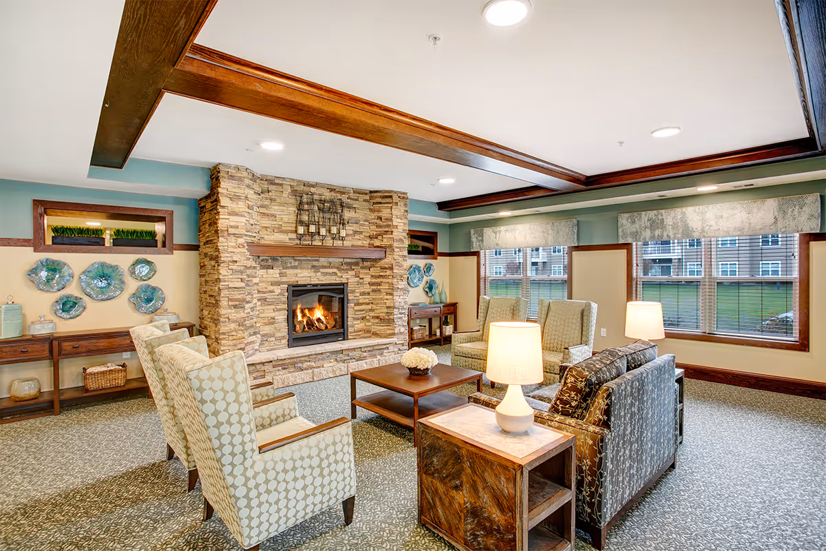 Well-lit senior living lounge with a stone fireplace, upholstered chairs and sofas, a coffee table, and large windows.