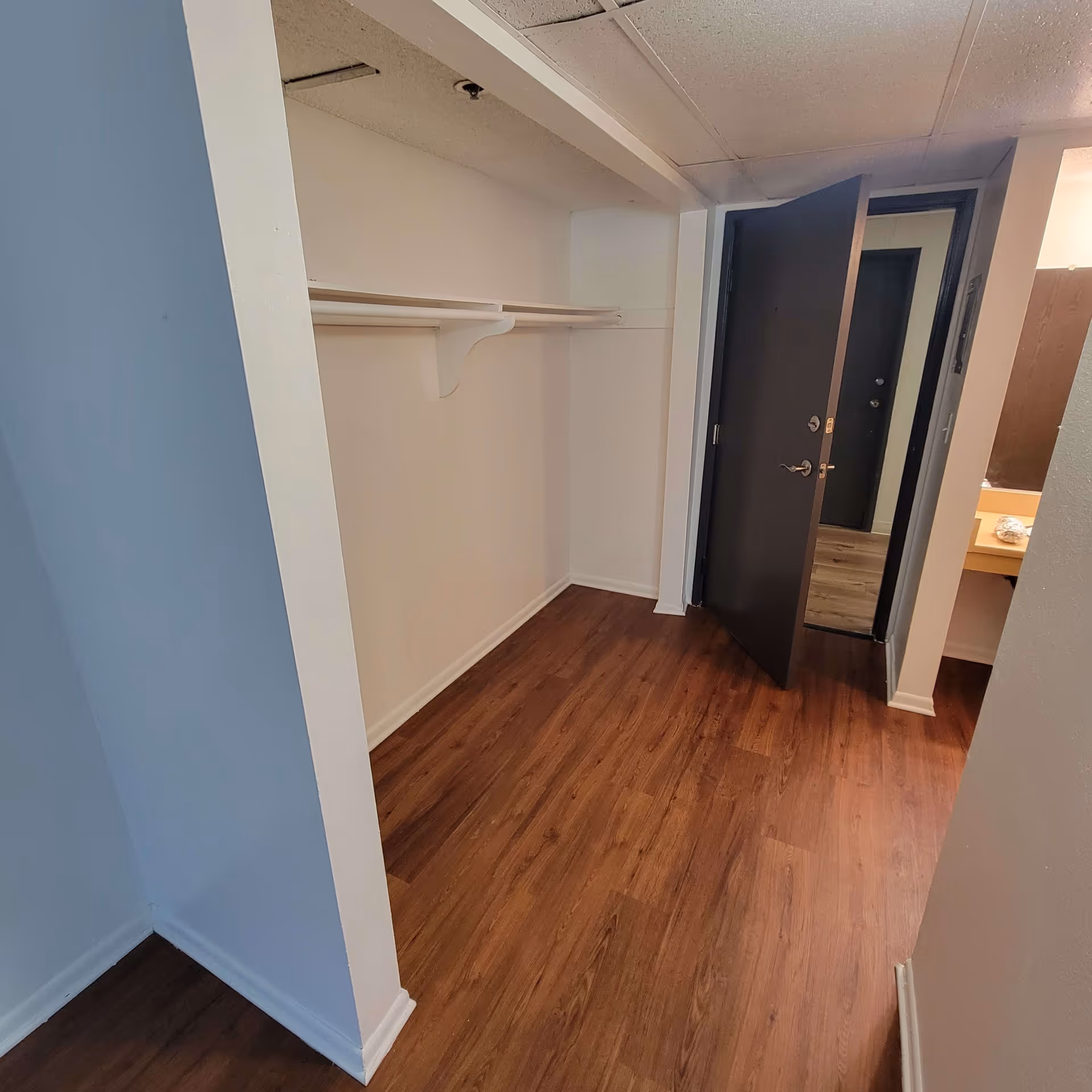 Empty walk-in closet with wooden floor, white walls, and a white shelf with hanging rods. A dark brown door is open, showing a glimpse of another room with similar flooring and a vanity area with a mirror and countertop.