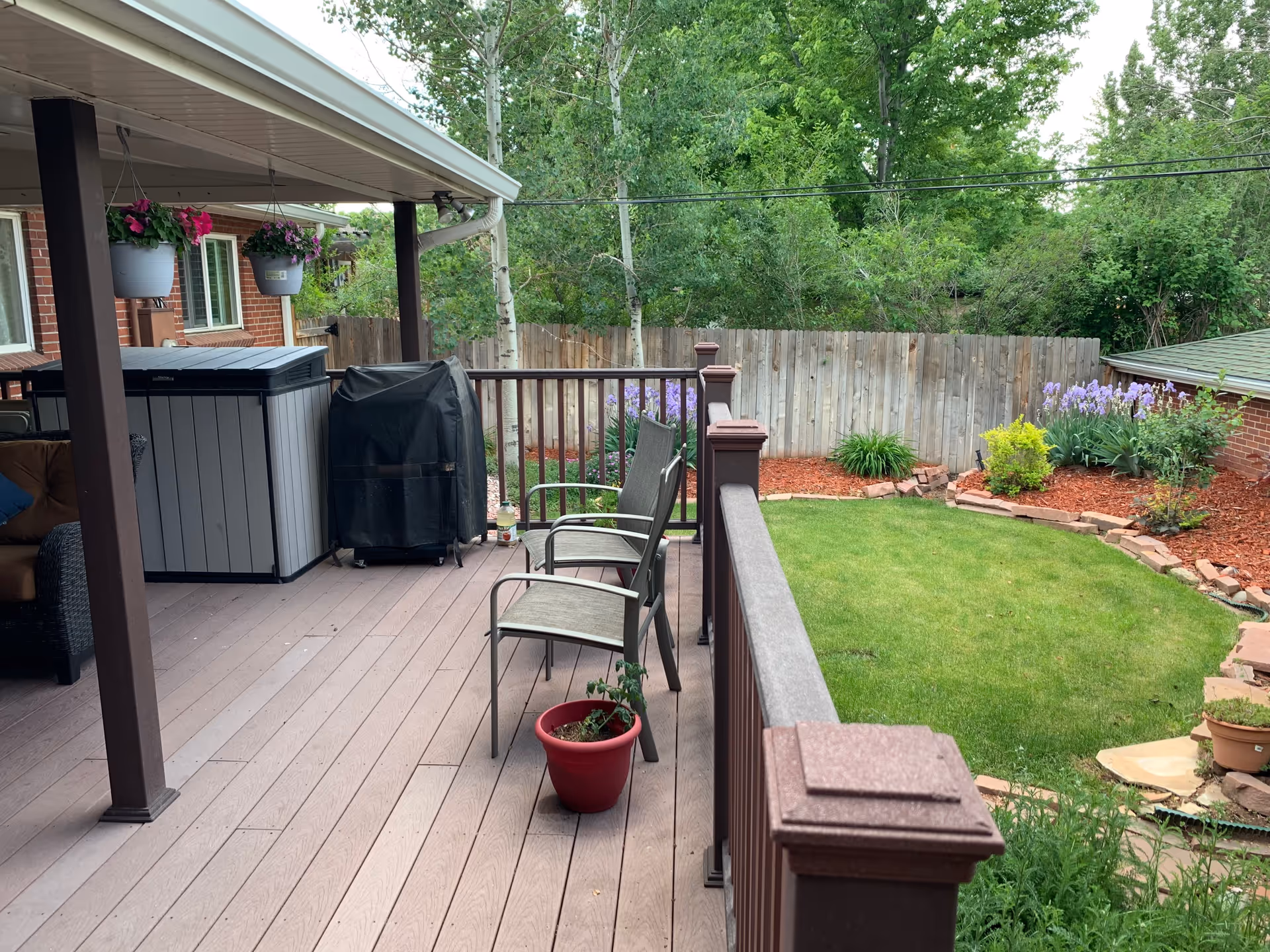 A covered outdoor patio area with two chairs, a small red potted plant, a covered grill, and a storage cabinet. The patio overlooks a well-maintained backyard with green grass, flower beds with purple and yellow flowers, and a wooden fence. Trees and shrubs are visible in the background.