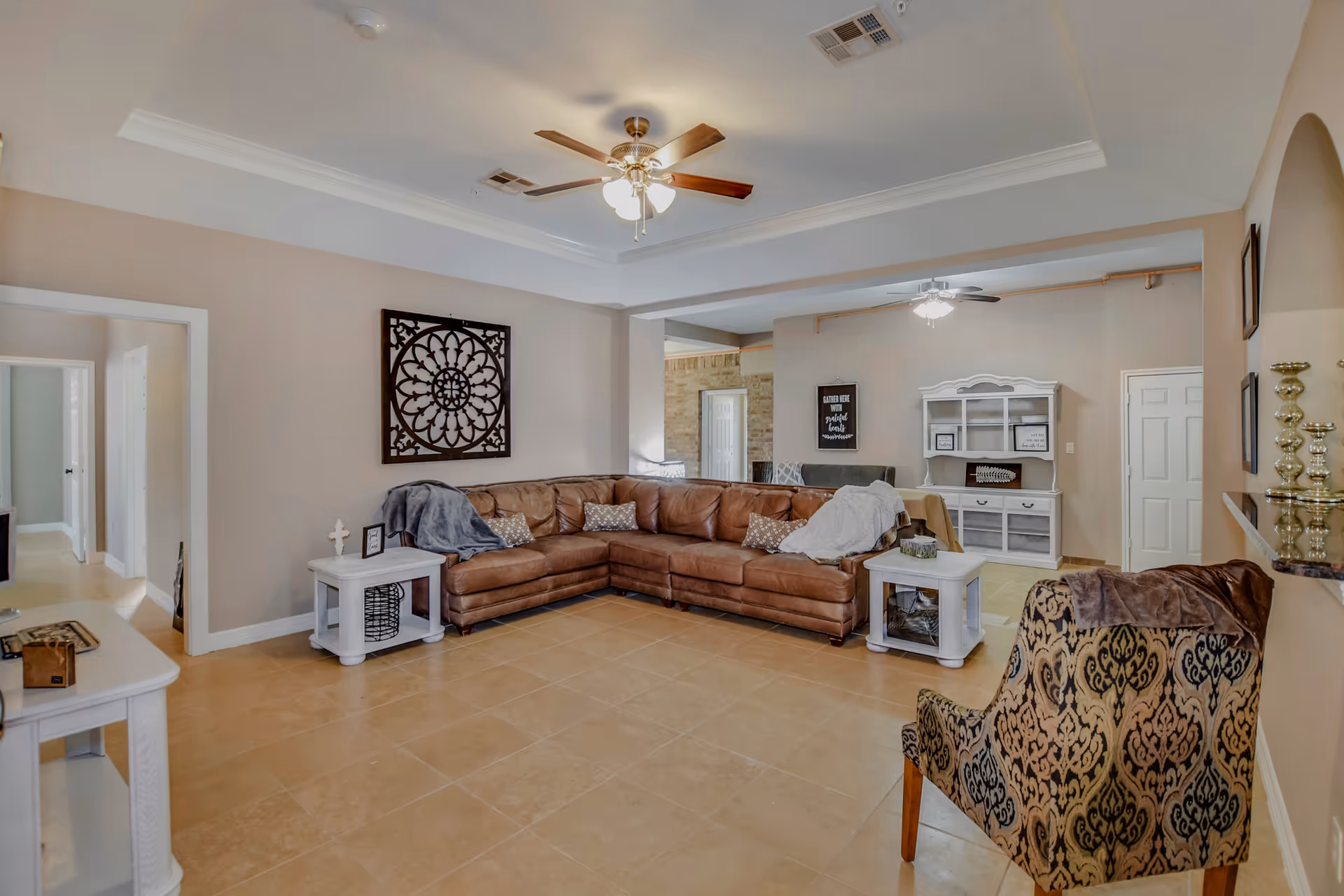 A spacious living room with beige tiled floors and light beige walls. The room features a large brown leather sectional sofa with pillows and throws, two white side tables, a patterned armchair, and ceiling fans with lights. Decorative wall art and shelves with ornaments are also visible.