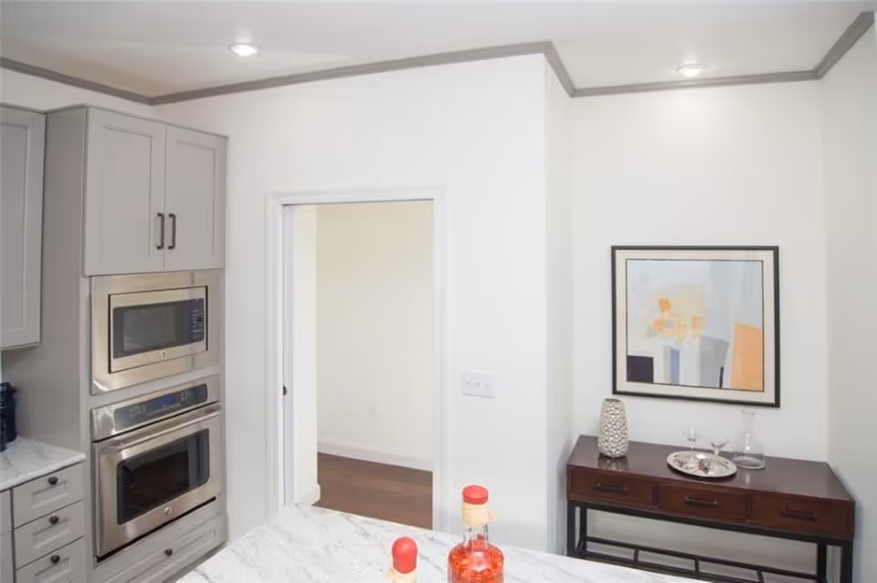 Modern kitchen interior with stainless steel built-in microwave and oven, gray cabinets, a marble countertop island with bottles, and a console table with artwork on the wall.