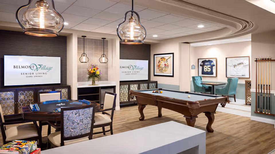 A well-lit senior living common area featuring a pool table with balls arranged on it, a card table with chairs, and two teal armchairs around a small table. The walls display framed sports jerseys and artwork. Two large screens show the Belmont Village Senior Living Sabre Springs logo. Pendant lights hang from the ceiling, and a vase with flowers is placed on a counter.