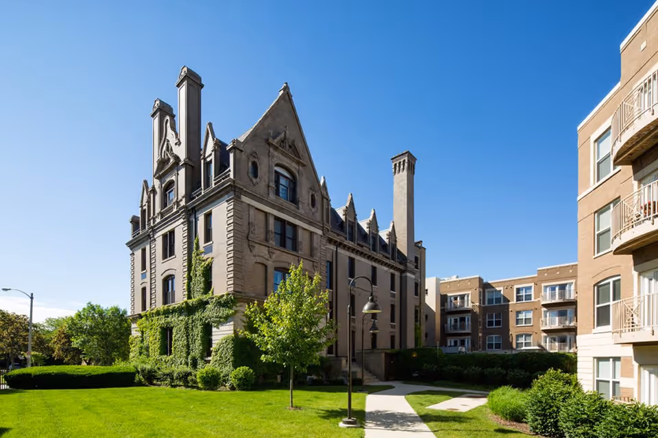 Exterior view of Eastcastle Place, a CCRC, showing a historic multi-story building with ivy growing on one side, surrounded by well-maintained green lawns, trees, and a modern apartment building under a clear blue sky.