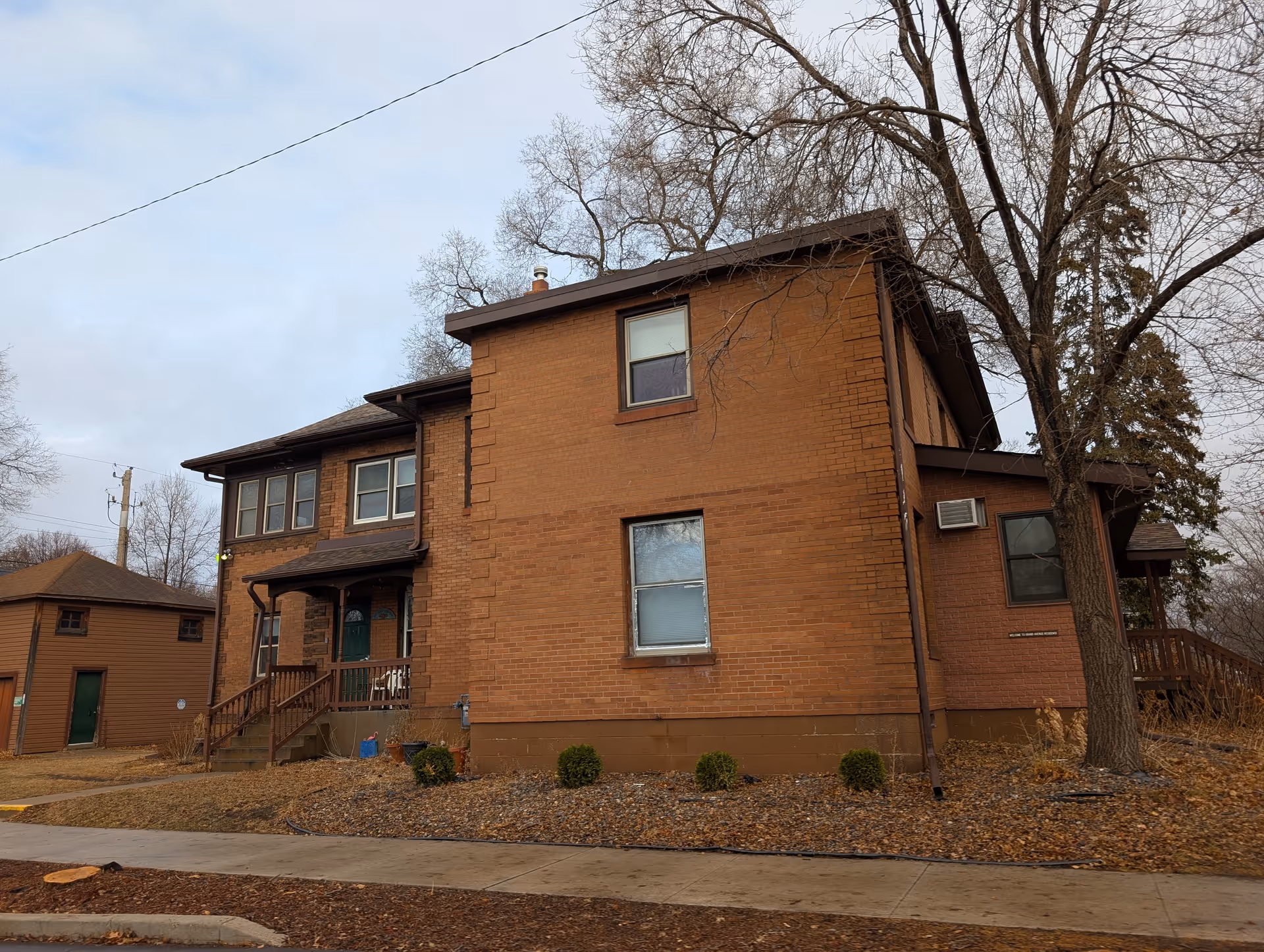 Exterior view of a two-story brick building with several windows and a small porch with stairs leading to a green door. There are leafless trees around the building and a sidewalk in front. The sky is overcast.