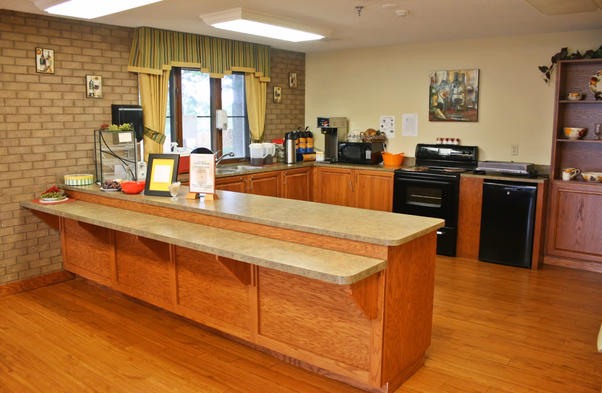 Communal kitchen with a long wooden island and countertop, sink, coffee station, stove and cabinets.