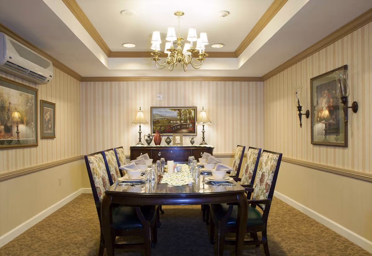 A formal dining room with a long wooden table set for eight people. The chairs have floral upholstery. The table is set with plates, glasses, and folded napkins. The room has beige striped wallpaper, a chandelier hanging from the ceiling, two table lamps on a sideboard, and framed paintings on the walls.
