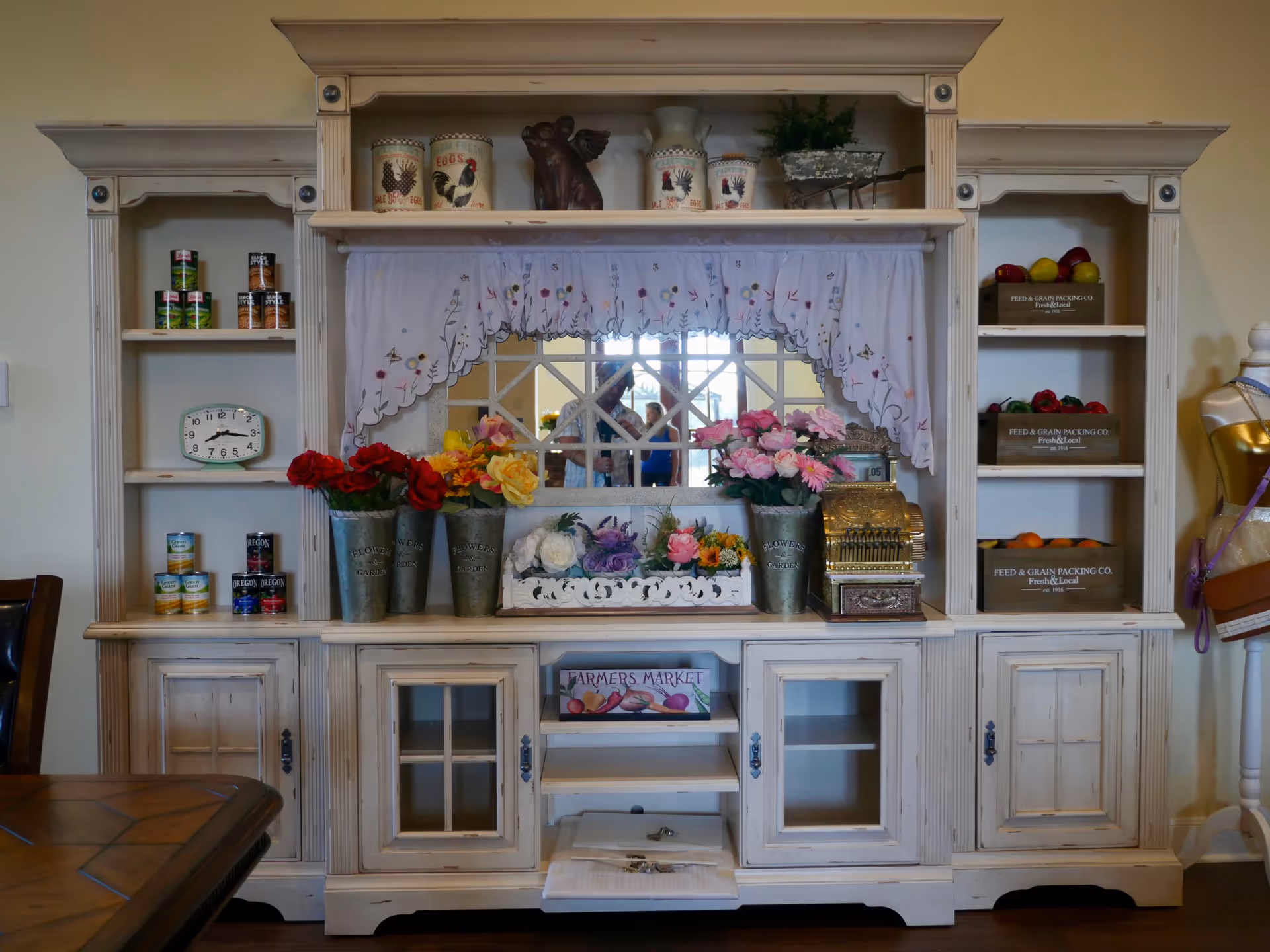 A rustic white wooden cabinet with shelves displaying decorative items including metal buckets with artificial flowers, canned goods, a vintage clock, wooden crates with artificial fruits, and a vintage cash register. A mirror with a white embroidered curtain is centered above the cabinet, reflecting part of the room and two people in the background.