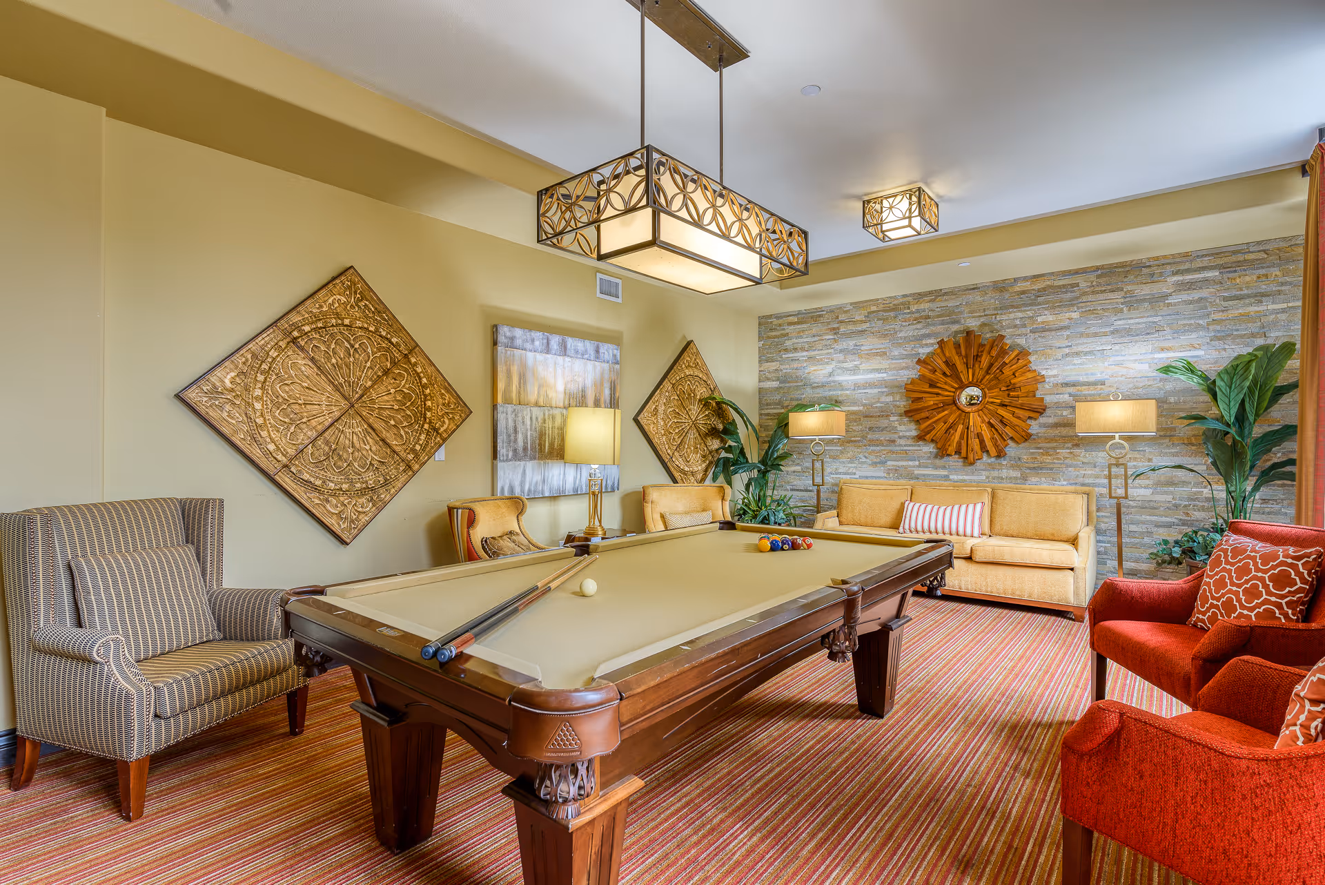 A cozy game room featuring a pool table with cues and balls set up, surrounded by comfortable seating including striped armchairs, yellow sofa, and red armchairs. The room has warm yellow walls, decorative wall art, a stone accent wall with a sunburst mirror, potted plants, and stylish hanging and floor lamps. The floor is covered with a striped carpet.