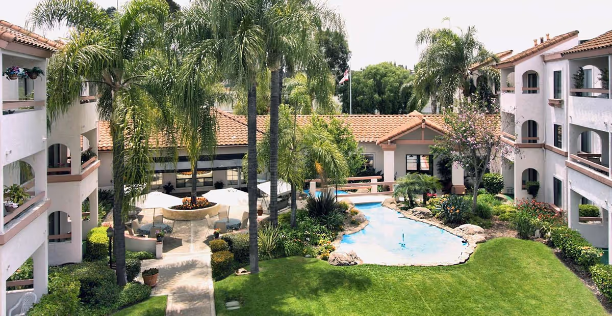 Sunlit courtyard with palm trees, a small pond or pool, umbrellas and seating surrounded by two-story white buildings with tiled roofs.
