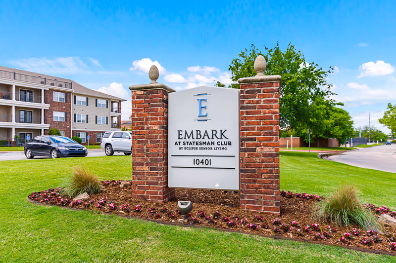 A brick and white sign for Embark at Statesman Club by Eclipse Senior Living, located on a well-maintained grassy area with flowers and landscaping. In the background, there is a multi-story residential building and parked cars under a blue sky with some clouds.