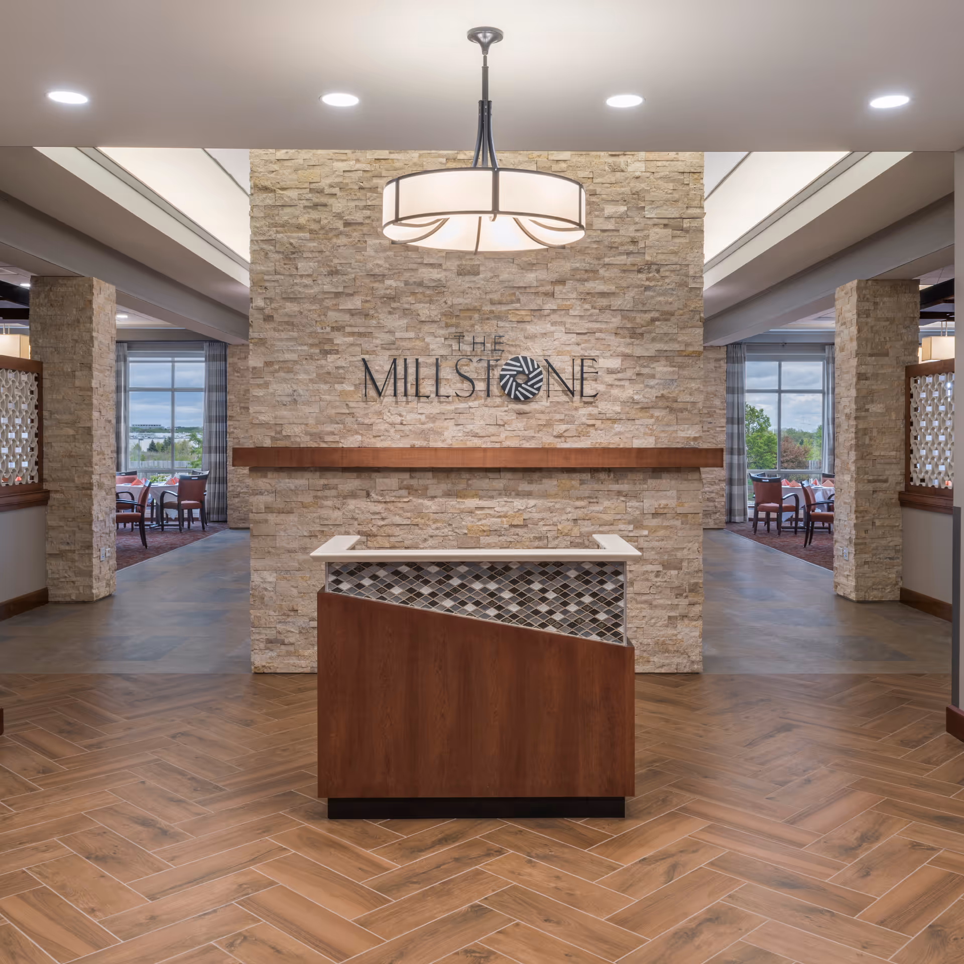 Interior view of a senior living facility lobby with a stone accent wall featuring the sign 'The Millstone'. A modern chandelier hangs from the ceiling above a wooden reception desk with a tiled front. The space has wood-patterned flooring and leads to dining areas with large windows and chairs visible on both sides.