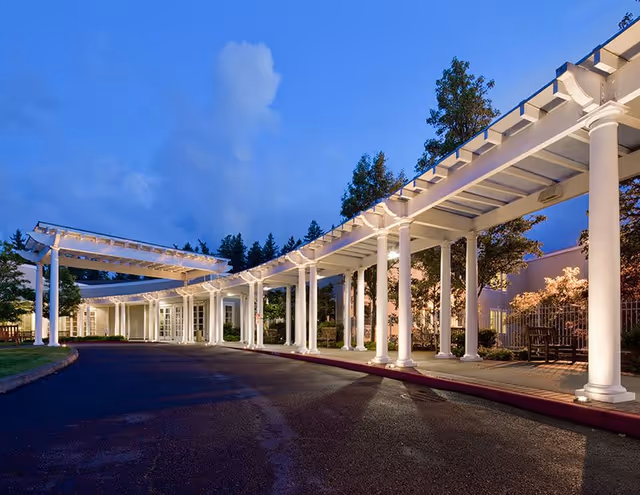 Evening view of the exterior entrance of The Rose Schnitzer Manor at Cedar Sinai Park, featuring a covered walkway with white columns and soft lighting, surrounded by trees and landscaping.