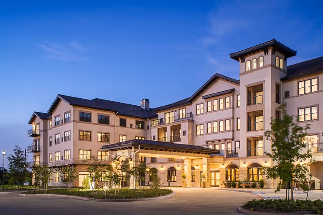 Exterior view of a large, multi-story senior living facility named Grand Living At Riverstone during twilight, with warm lights glowing from the windows, a covered entrance, and landscaped greenery around the driveway.