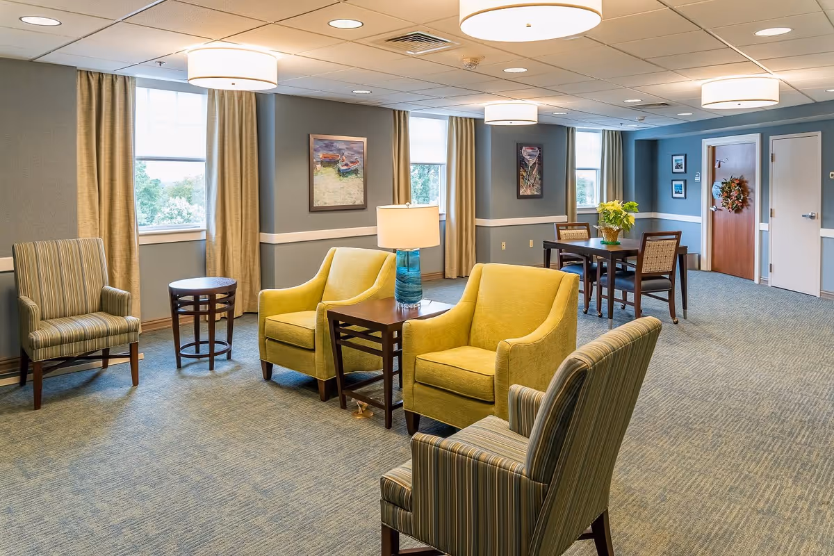 A bright and spacious common area in a senior living facility featuring two yellow armchairs and two striped armchairs arranged around a wooden side table with a blue glass lamp. There is a small round table near the window and a dining table with four chairs in the background. The walls are painted blue with white trim, and there are several windows with beige curtains letting in natural light. Artwork is hung on the walls, and the carpet is a muted blue pattern.