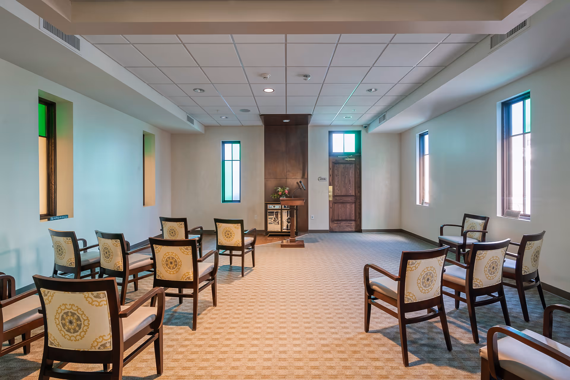 A small interior meeting/chapel-style room with rows of wooden chairs facing a podium and a door, lit by narrow windows.