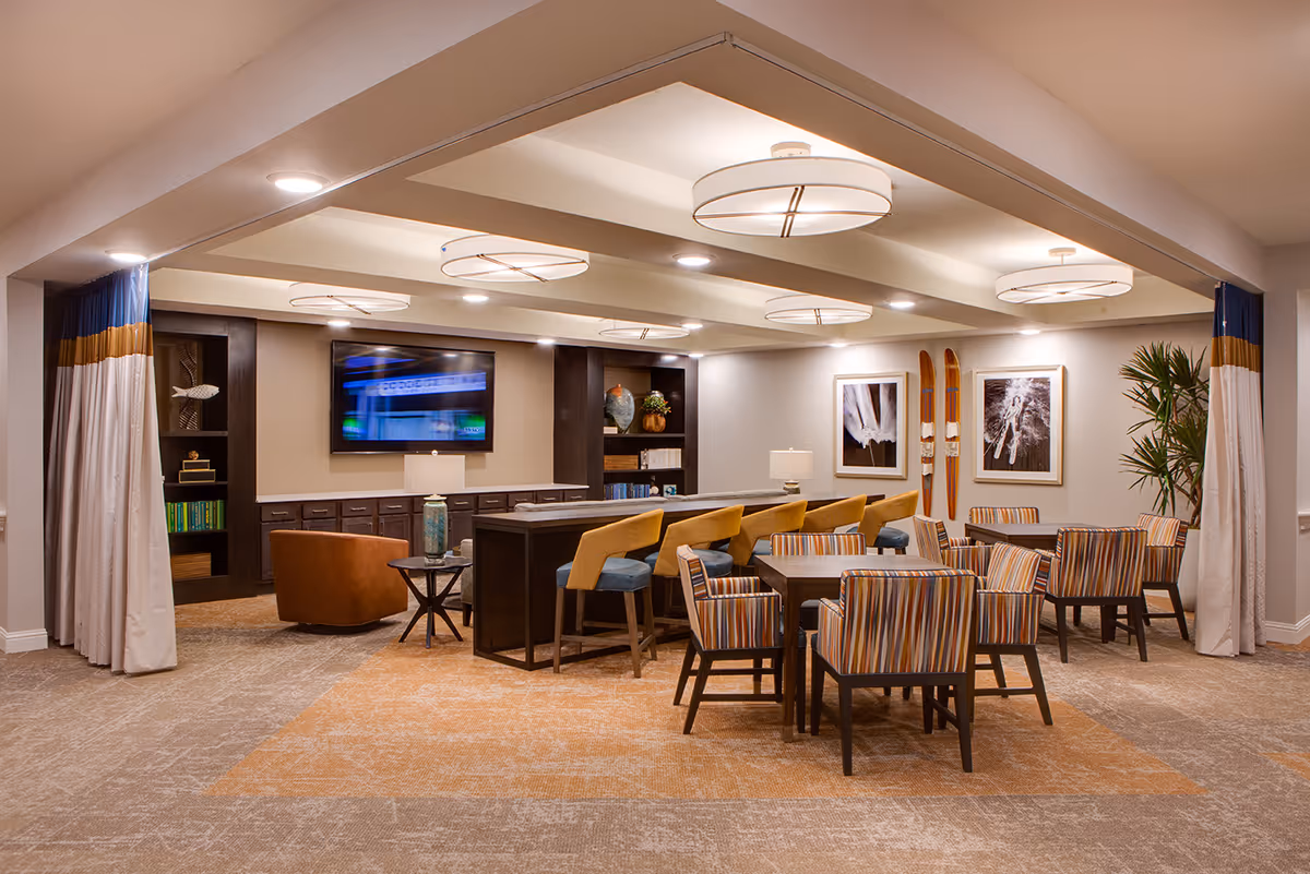 A spacious and well-lit common area with a mix of seating options including a row of bar stools at a high counter, a round table with striped upholstered chairs, and a single brown armchair. The room features built-in dark wood shelving with decorative items and books, a wall-mounted flat-screen TV, modern ceiling lights, and framed artwork on the walls. There are also curtains with blue and gold accents framing the entrance to the room.
