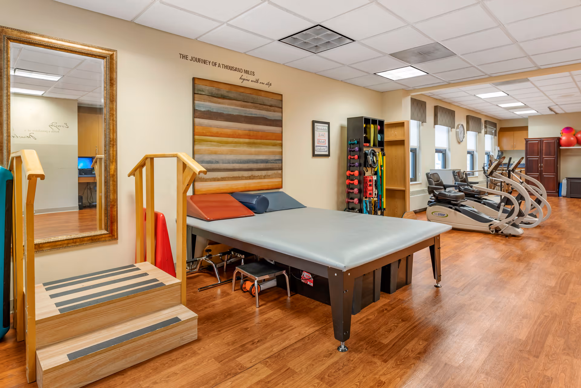 A rehabilitation or physical therapy room with exercise equipment including recumbent bikes, a padded therapy table with cushions, a small set of wooden stairs with handrails, and a rack holding various exercise weights and bands. The room has wood flooring, multiple windows with blinds, and motivational wall art that reads 'The journey of a thousand miles begins with one step.'