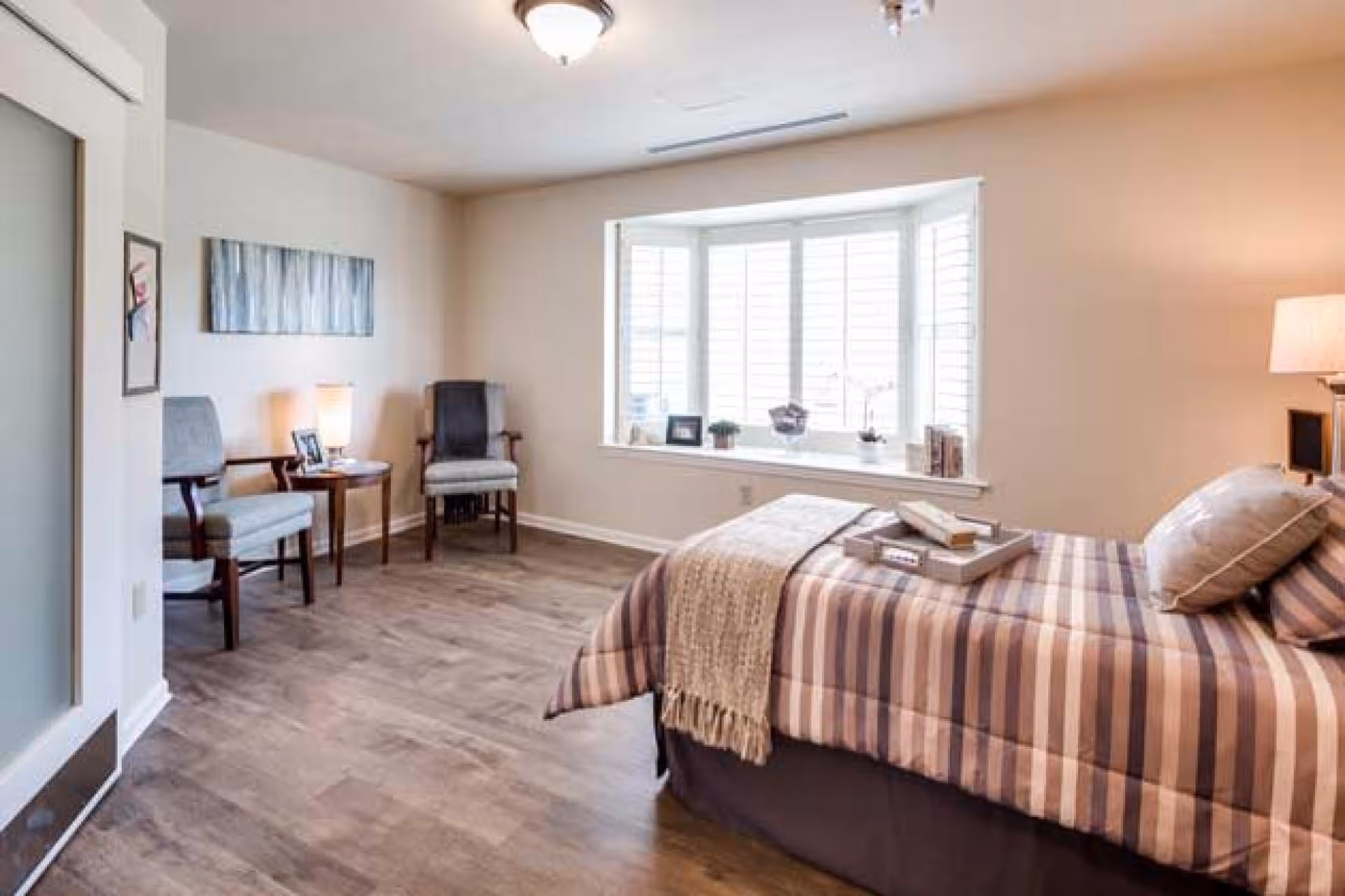 Sunlit bedroom with a striped bed, tray, two chairs and a side table, and a large bay window.