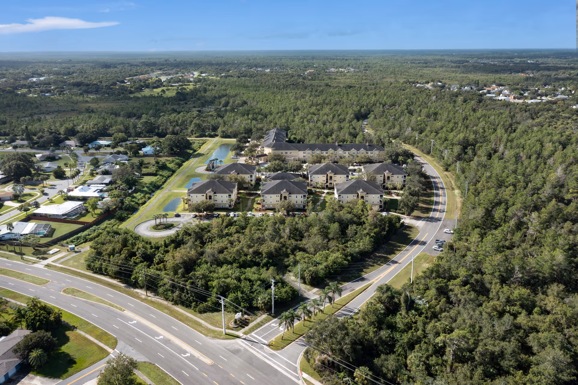 Aerial view of Glenbrooke Senior Living facility surrounded by dense greenery and trees, with multiple beige buildings arranged in a campus layout. There are roads and parking areas around the buildings, and a small pond with a gazebo is visible within the property. The surrounding area includes residential houses and forested land extending to the horizon under a clear blue sky.