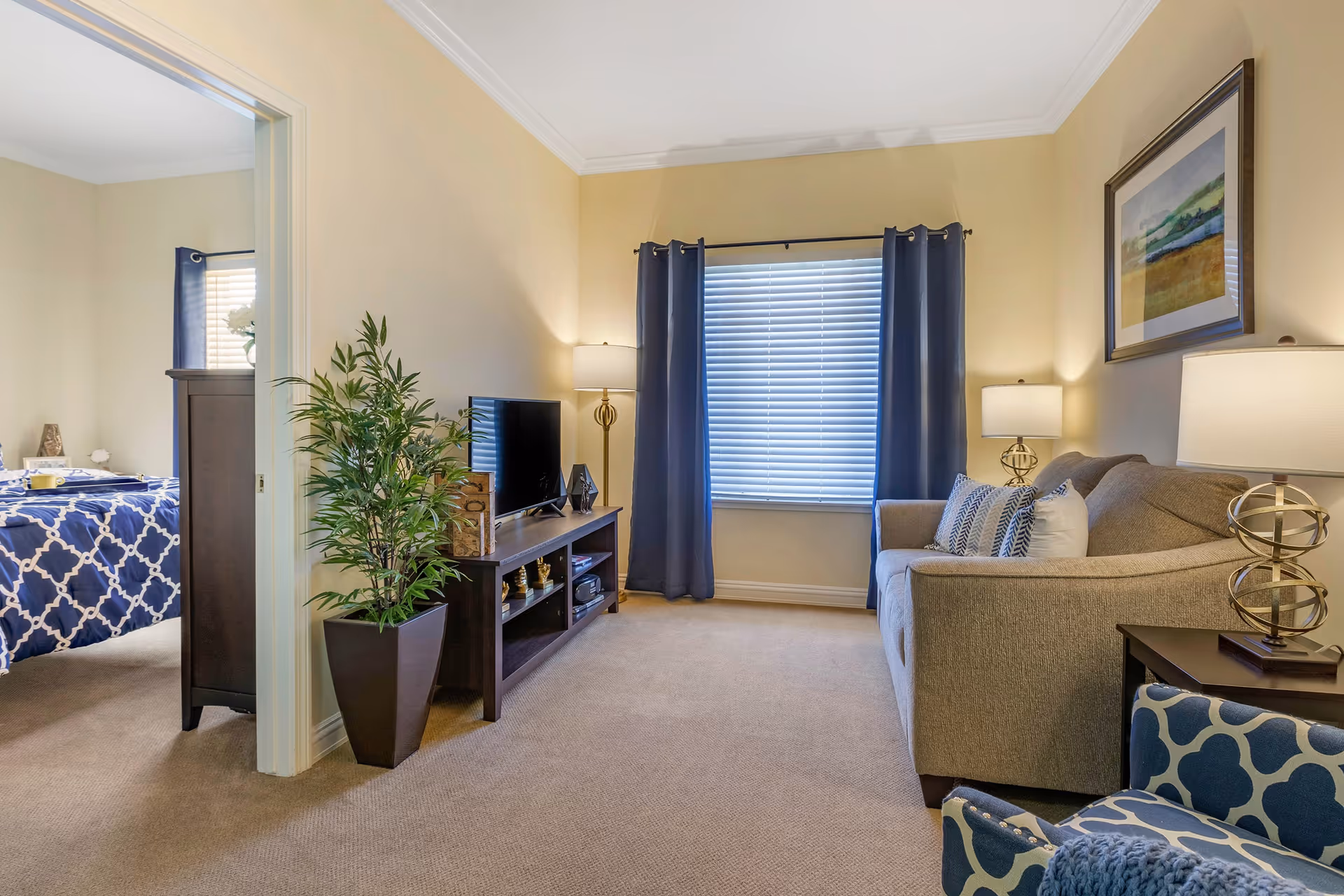 A cozy living room with beige walls and carpet, featuring a gray sofa with patterned pillows, a blue patterned armchair, two modern lamps, a TV on a wooden stand, and a large window with blue curtains. To the left, an open doorway reveals a bedroom with a bed covered in a blue and white patterned comforter.