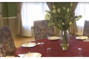 A round dining table with a red tablecloth set with plates and glasses and a vase of tulips, surrounded by upholstered chairs in a bright room with curtained windows.