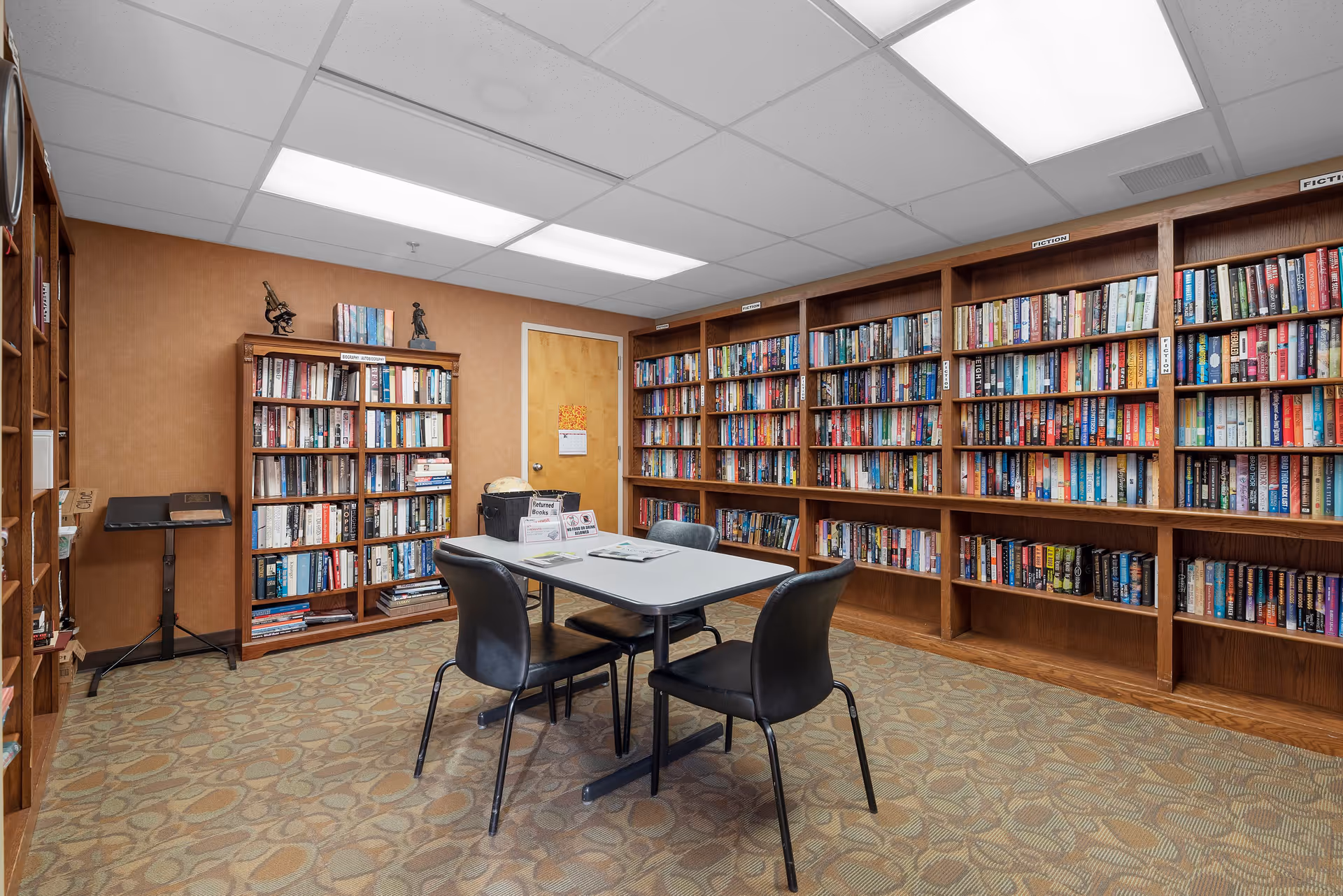 A cozy library room in a retirement community with wooden bookshelves filled with books along two walls. In the center, there is a rectangular table with four black chairs around it. The room has a patterned carpet, a closed wooden door, and fluorescent ceiling lights.